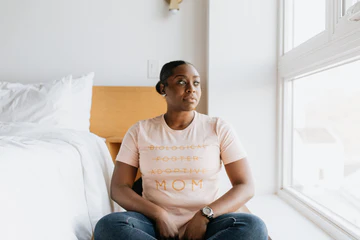 Black woman wearing G&B shirt that says Biological, Adoptive and Foster all crossed out but the word MOM is not. She is sitting on a stool besides a bed looking out a window.